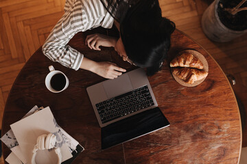 Dark-haired girl fell asleep on wooden table while working in laptop. There is cup of coffee and croissants on table