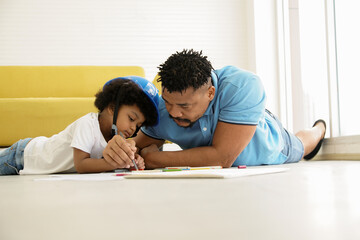 African American family father and son drawing and lay on the floor at home. Young father holding his little boy hand, holding a pencil to teach drawing and painting on paper