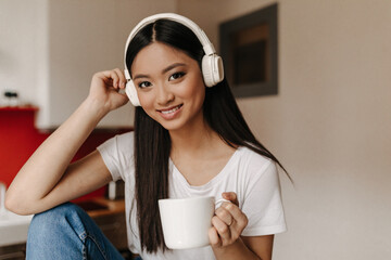 Brunette woman with charming smile looks into camera, holding big cup. Asian listens to music on headphones