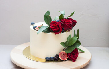 Elegant wedding cake decorated with fresh red roses, green leaves, figs and blueberry. Plain grey background.