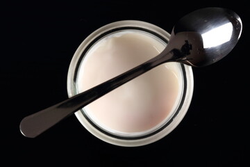 Yogurt in open small glass jar with spoon at black background. D