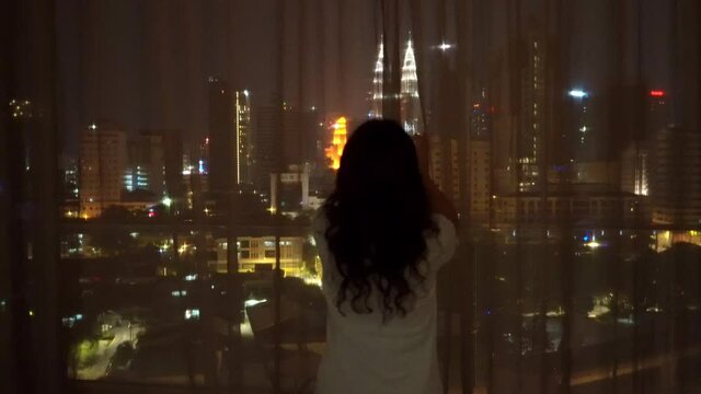 The girl opens the curtains and opens a breathtaking view of the large night city with many lights in front of her. Evening view of the Petronas Towers 