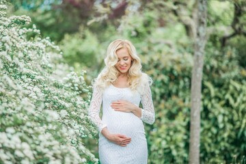 Naklejka premium Young beautiful pregnant woman in a lace white dress walks in a flowering park. Portrait of a beautiful pregnant blonde. Spring.