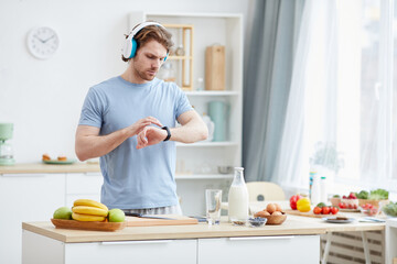 Young man wearing headphones and checking time on his watch while cooking breakfast in the kitchen