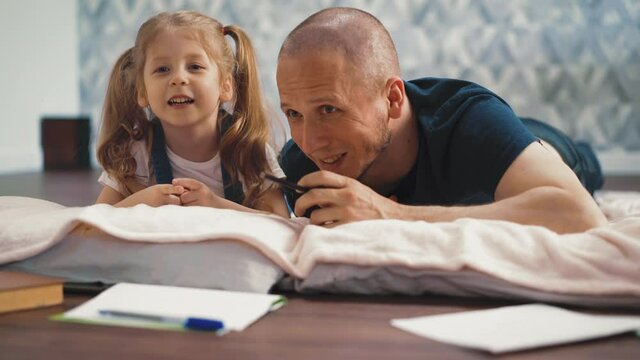 close-up little daughter with bald dad lie on the floor, hiding, looking into something forward, at each other, laughing, smiling, talking, around school accessories, paper