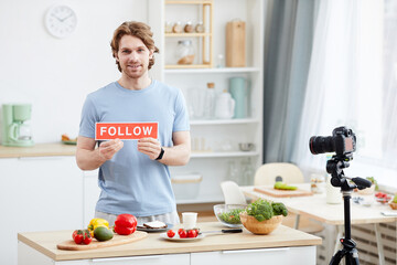 Portrait of young man with placard Follow smiling at camera and shooting blog for his followers in domestic kitchen