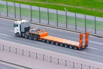 Truck with a trailer and an empty orange long platform rides in the city on the highway.