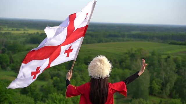 Georgian girl with national flag of Georgia in hands walks around green hills. Georgian culture lifestyle. Woman in papakha and red dress