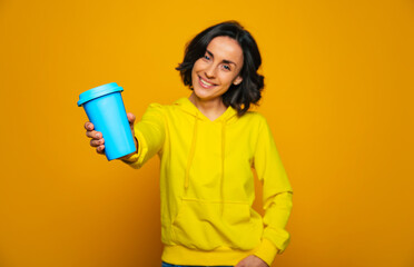 Try my drink. Half-length photo focused on thermal mug, holded on outstretched arm by attractive woman, dressed in a yellow hoodie, offering to try her drink.