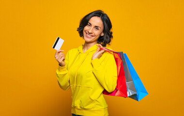 Thanks to my credit card! Half-length photo of a young girl, dressed in a yellow hoodie turned sideways, smiling at the camera, showing her credit card purchases in colorful bags.