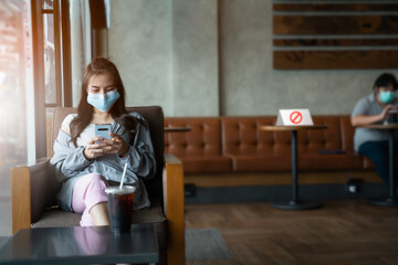 Social distancing concept.Young asian woman in facial mask sitting use social media on the cafe and distance of 6 feet from other people keep distance protect from COVID-19 viruses at coffee cafe.