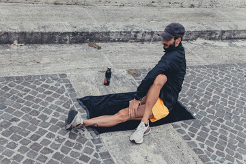 Athletic man practices sport and stretching exercises sitting on the ground alone on a city sidewalk