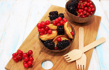 Tiny pancakes with berries, red currants and blackberries in coconut bowls and wooden forks on a  plate
