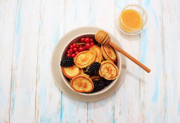 Tiny pancakes with berries of red currant, blackberry, honey in a beige bow and wooden spoonsl on a wooden background