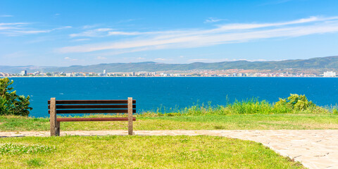 Obraz premium bench on the sunny beach shore. beautiful view from paved footpath on the seaside. city and mountain in the distance beneath a blue sky with clouds