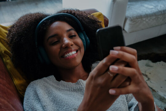 Beautiful Young Woman With Earphones On And Enjoying Downtime On The Couch With Her Cell Phone, Browsing The Internet And Social Media