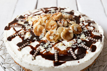 frisch gebackene Torte mit Bananen, Sahne, Schokosoße, Karamellsoße und Haselnussboden auf Tisch aus Holz in der Küche zum Kaffeetrinken