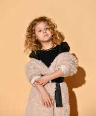 Pretty blonde curly model in necklace, black dress and faux fur coat with belt. She is smiling, posing against beige background