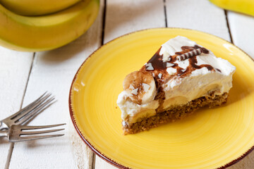 frisch gebackene Torte mit Bananen, Sahne, Schokosoße, Karamellsoße und Haselnussboden auf Tisch aus Holz in der Küche zum Kaffeetrinken