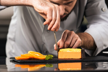 Obraz premium Chef male decorate cake at kitchen. Close up chef hands make orange cake.