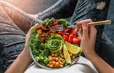 Woman in jeans holding Buddha bowl with salad, baked sweet potatoes, chickpeas, broccoli, greens, avocado, sprouts in hands. Healthy vegan food, clean eating, dieting, top view, toning