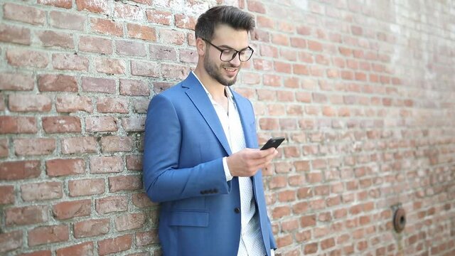 businessman leaning on a brick wall, texting on his phone, smiling and being surprised