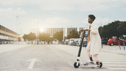 Young fancy African woman in a white trench is using e-scooter on the road with an arrow marking on it; a charming black girl in a beige cloak and spectacles is riding an electric scooter outdoors