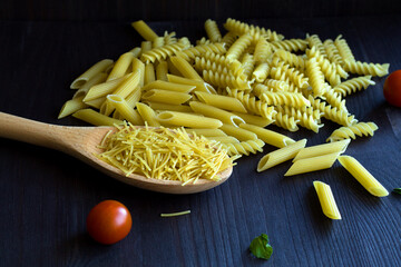 raw macaroni with fusilli, penne with cherry tomatoes on a black background. different types of Italian pasta with a wooden spoon on a table with copy space.