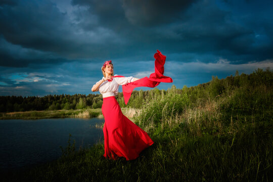 A slender caucasian girl in a red skirt and white blouse is dancing with a red shawl in her hands against a stormy sky
