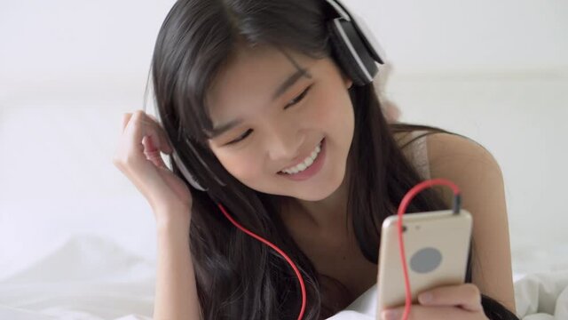 Beautiful young asian woman lying happy earphone listening music with relax and enjoy in the bedroom, girl fun and relax headphone with playing song mp3 on smart mobile phone, lifestyle concept.