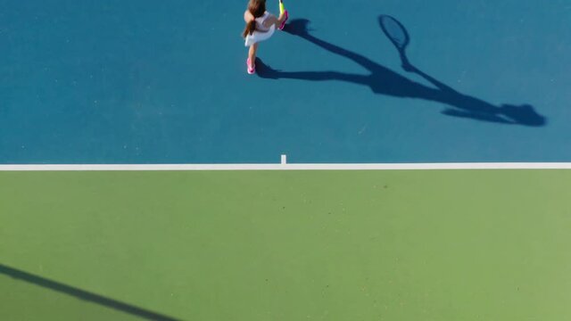 4K aerial top-down view of a young female sportsman during her practice. A close-up of a girl athlete serves the tennis ball. Young woman is hitting the ball with her tennis racket at sunset