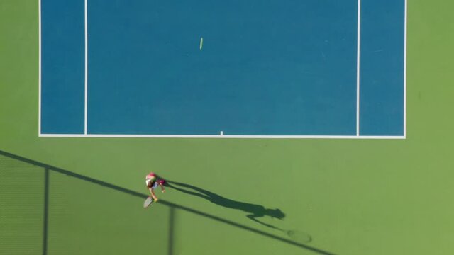 An energetic young woman is actively playing tennis by and hitting the ball with her racket. 4K aerial top-down view of a female athlete during her practice. Sport activity on a sunny day.