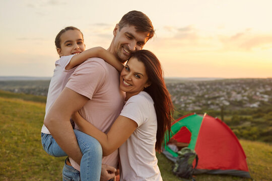 Happy Young Parents With Child Hugging At Their Camp In Mountains. Girl With Mom And Dad Embracing On Camping Trip