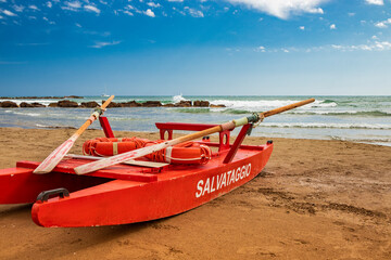 Typical red rescue boat, with oars, used by Italian lifeguards, stationary on the sand. "Rescue" writing on the side. Blue sky with white clouds in summer on the Roman coast. Wind and rough sea.