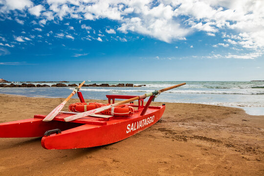 Typical red rescue boat, with oars, used by Italian lifeguards, stationary on the sand. "Rescue" writing on the side. Blue sky with white clouds in summer on the Roman coast. Wind and rough sea.