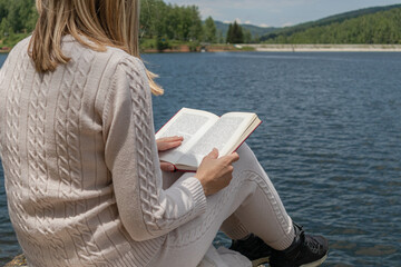 Girl reading a book near to lake water in a sweater on a sunny day. Blonde girl sitting on a large rock on the shore of the lake with a book on legs. Relaxation concept. Close up, selective focus