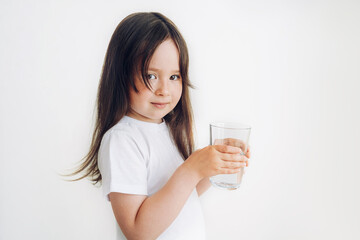 A child in a white T-shirt drinks water. Little girl stands with a glass of water