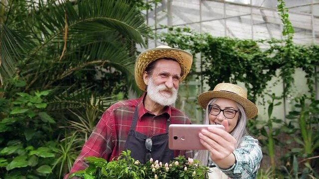 Likable positive senior couple in straw hats making funny grimaces while making photos in beautiful glasshouse