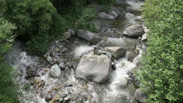 View from above of a beautiful mountain stream: it is laden with water that creates white foam beating among the gray rocks. The vegetation runs alongside the stream.