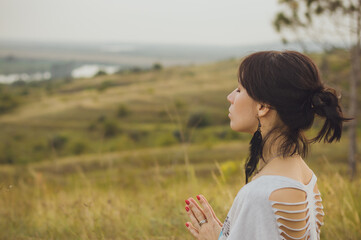 A dark-haired girl in a white t-shirt meditates, practicing yoga. Hands in the Namaste position from behind. Nature, beautiful landscape, autumn.