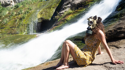 Woman with gas mask and yellow dress at a waterfall in nature. There is no planet b