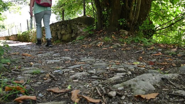 Imagine close to the ground of a path: you can see the boots of a woman who is walking with a red backpack on her shoulders.