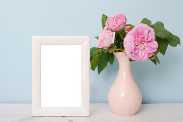 Mock-up empty photo frame near a vase with flowers on a table on blue wall background