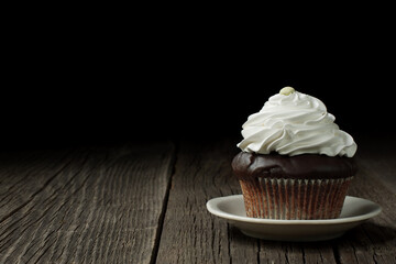 A chocolate cupcake in a typical fluted cupcake liner, topped with whipped cream and decorated with a button-shaped candy.  It is standing on a small ceramic saucer, on rustic weathered wood planks. 