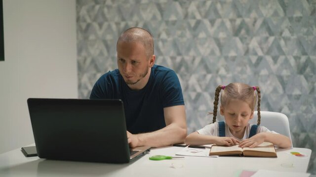 concept of father and daughter home learning: little girl reads book aloud, leads her index finger on lines, dad sits next to table and works, communicates with colleagues via video link on computer