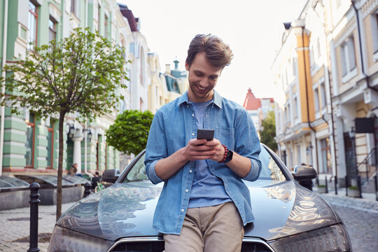 Delighted Young Male Sitting On Car Hood With Cell Phone