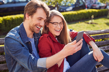 Cheerful man and woman resting on bench with a smartphone
