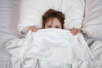 Small boy lies in bed with his eyes open, afraid of nightmares and terrible dreams in children