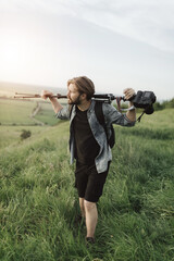 Fototapeta premium Tired bearded man in summer outfit holding tripod with digital camera on shoulder while climbing on high hill. Concept of professional photography outdoors