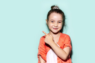 Studio portrait of smiling little child girl pointing with finger on empty background of aqua menthe color with copy space. Wearing coral pink shirt.